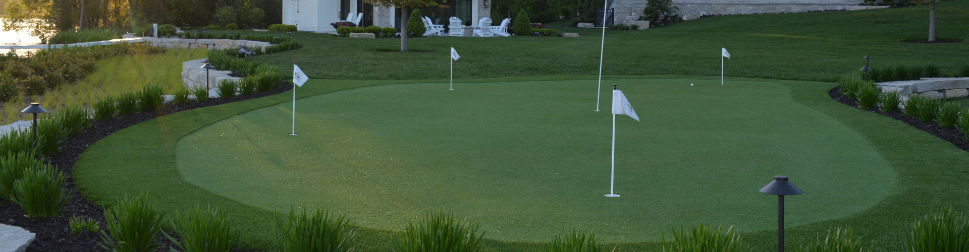 Putting green with four white flag pins surrounded by ornamental grass borders, landscape lighting, and a luxury estate with a pool house at sunset